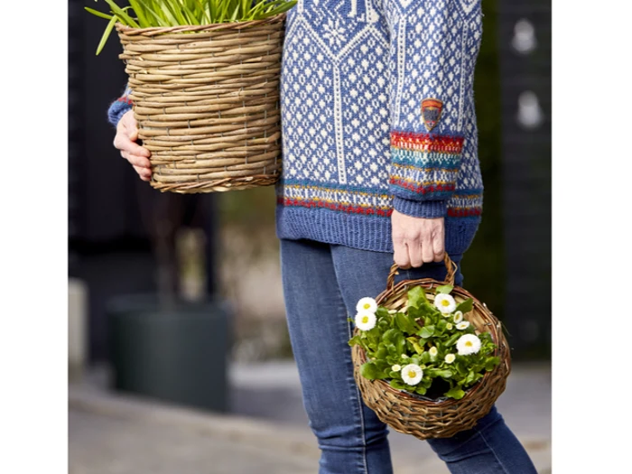 Person i blåmønstret sweater med en stor kurv med grønne planter og en lille flettet kurv med hvide margueritblomster.