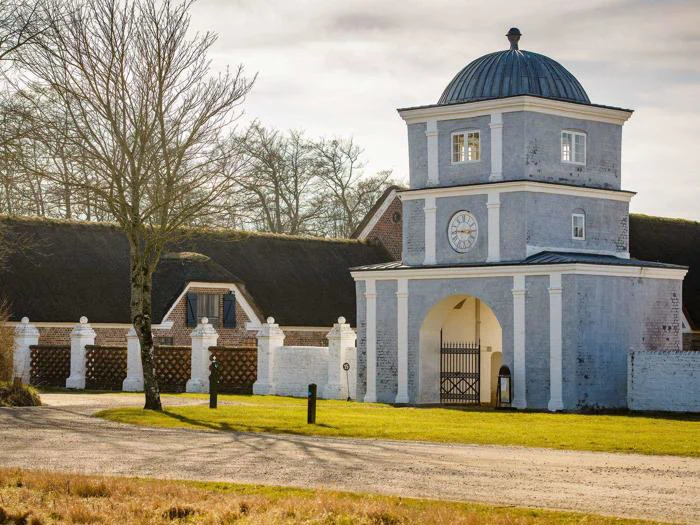 En lyseblå murstensbygning med klokketårn og buet indgang står nær en stråtækt bygning, et hvidt hegn og et bladløst træ på en solrig dag.