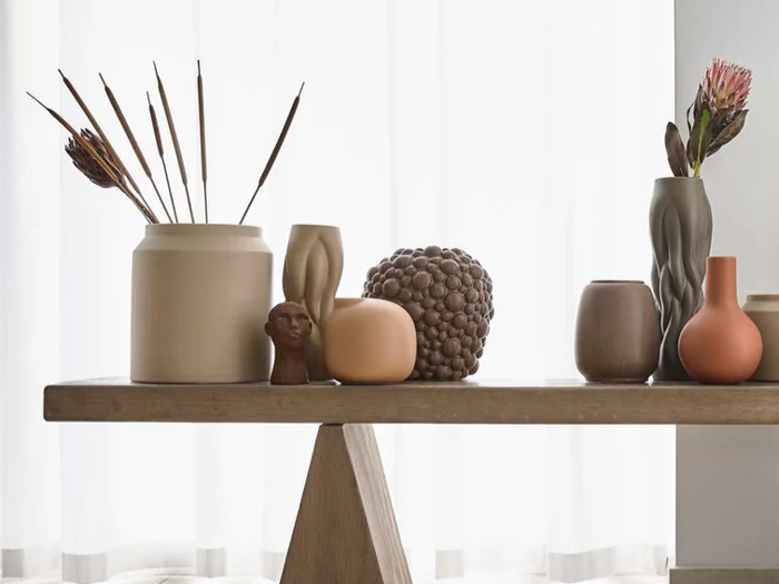 På et træbord står forskellige neutralfarvede keramikvaser, et skulpturelt hoved, tørrede planter og et rundt objekt med tekstur foran hvide, gennemsigtige gardiner.
