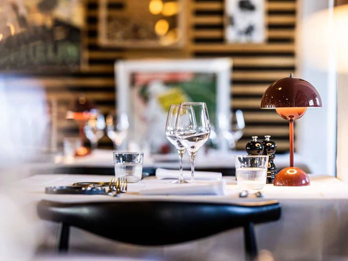 Nærbillede af et restaurantbord dækket med vinglas, vandglas, tallerkener, bestik, en rød lampe og salt- og peberbøsser med abstrakt kunst på væggen i baggrunden.