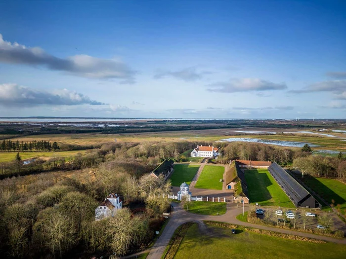 Luftfoto af et landligt landskab med spredte huse, grønne marker, træer og vand i det fjerne under en blå himmel med skyer.