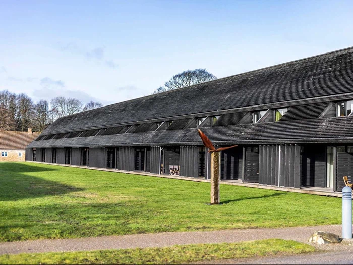 En moderne sort træbygning med skråt tag og flere vinduer, der ligger ved siden af et stort græsareal og en skulptur i forgrunden.