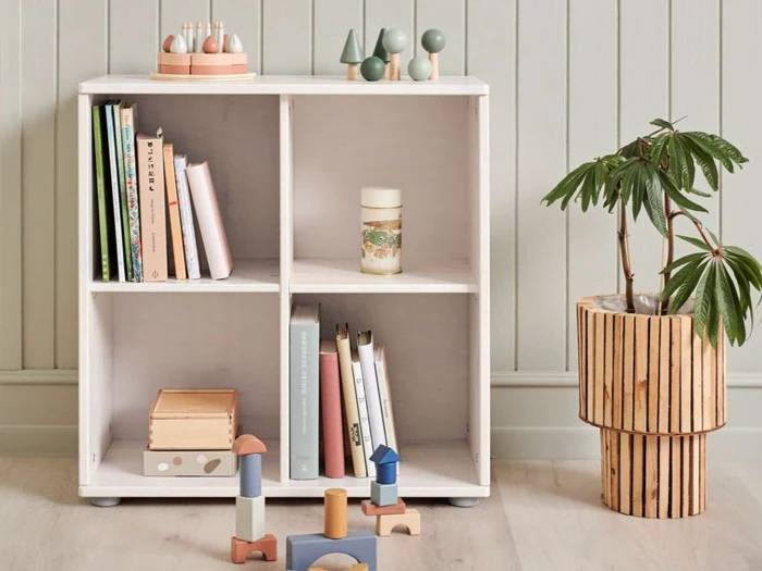 Hvid hylde med bøger, trælegetøj og kunstartikler ved siden af en potteplante i en træholder på et lyst trægulv mod en lys panelvæg.