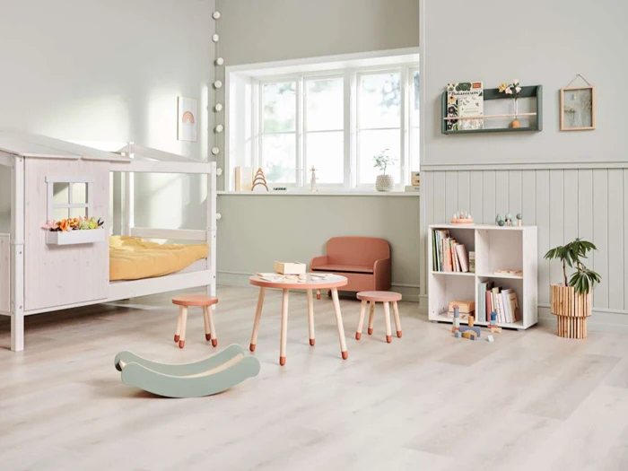 Et lyst, moderne børneværelse med en seng formet som et hus, et rundt bord med taburetter, bogreoler, legetøj, en potteplante og et stort vindue, der lukker naturligt lys ind.