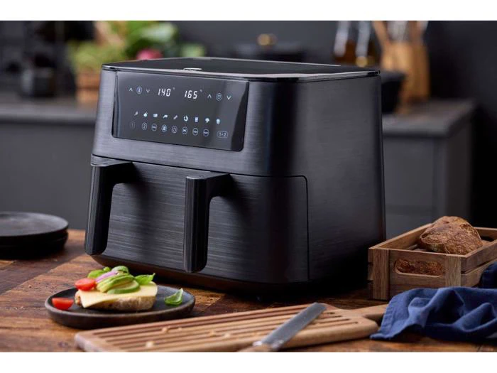 En sort frituregryde med to kurve og et digitalt display står på et køkkenbord af træ ved siden af en tallerken med mad, brød og køkkenredskaber.