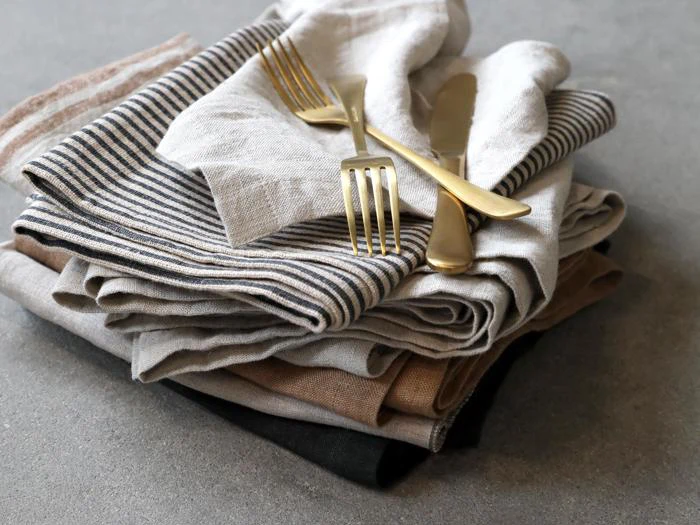 En stak foldede linnedservietter i neutrale toner med striber, toppet med to guldfarvede gafler og en kniv på en grå overflade.