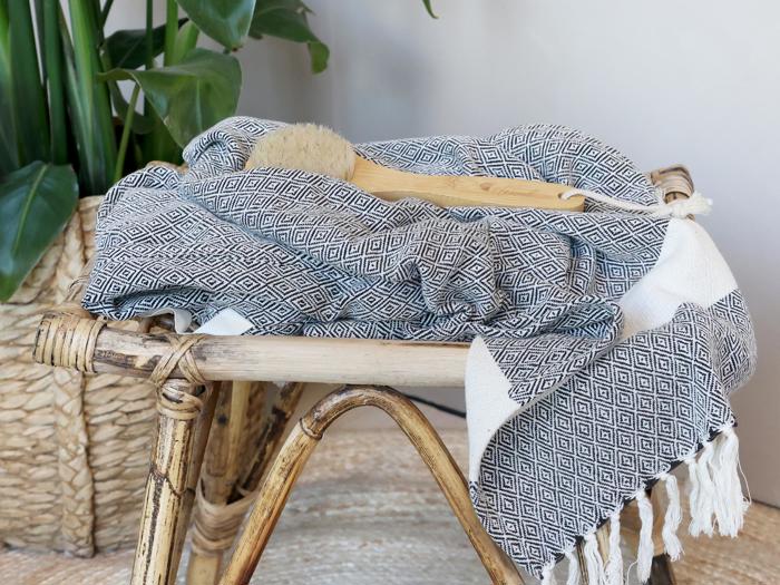 Et mønstret tæppe med frynsede kanter er draperet over en vævet bambusskammel ved siden af en naturbørste og grønne potteplanter i baggrunden.
