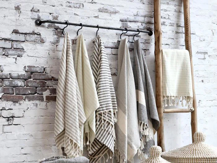Flere stribede og ensfarvede håndklæder hænger på kroge på en sort stang op ad en hvid murstensvæg med en træstige og flettede kurve i nærheden.