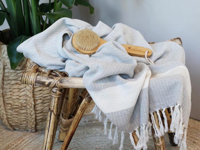 Et vævet håndklæde med geometrisk mønster og frynser er draperet over en rustik skammel med en badebørste af træ ovenpå. I baggrunden ses en flettet kurv med en plante.