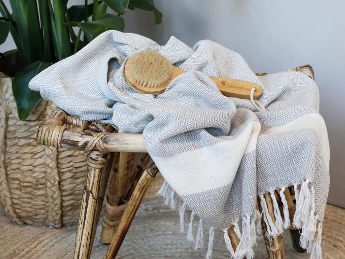Et vævet håndklæde med geometrisk mønster og frynser er draperet over en rustik skammel med en badebørste af træ ovenpå. I baggrunden ses en flettet kurv med en plante.