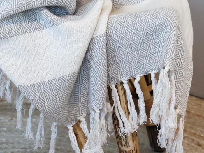 Et nærbillede af et gråt og hvidt vævet tæppe med diamantmønster og frynser, draperet over en rustik træskammel.
