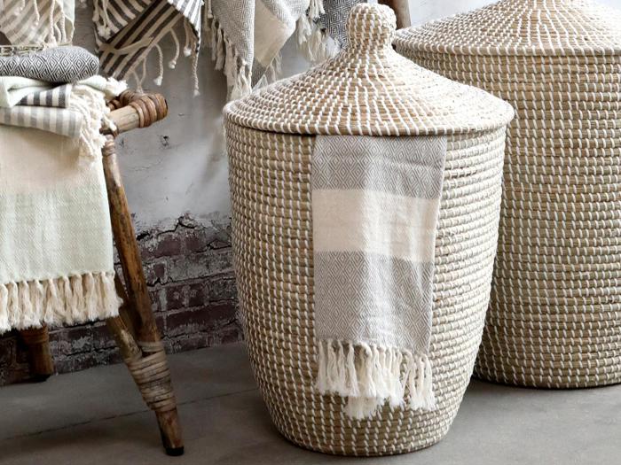 En flettet kurv med låg og et stribet tæppe med frynser draperet over siden står ved siden af en rustik træskammel med sammenfoldede tekstiler.