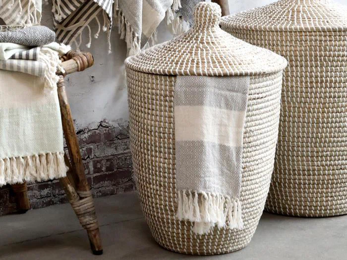 En flettet kurv med låg og et stribet tæppe med frynser draperet over siden står ved siden af en rustik træskammel med sammenfoldede tekstiler.