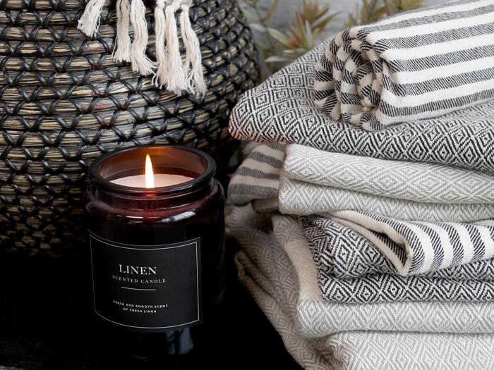 Et tændt linnedduftlys står ved siden af en stak mønstrede, foldede håndklæder og en vævet kurv med frynser i et hyggeligt miljø.