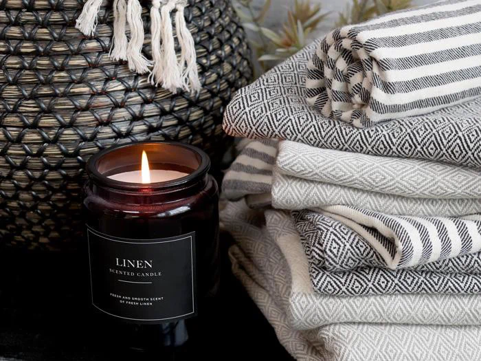 Et tændt linnedduftlys står ved siden af en stak mønstrede, foldede håndklæder og en vævet kurv med frynser i et hyggeligt miljø.