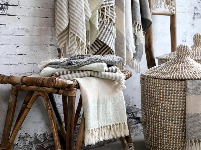 Flettede tæpper er draperet over en træbænk ved siden af to flettede kurve med låg, der står op ad en hvid murstensvæg.