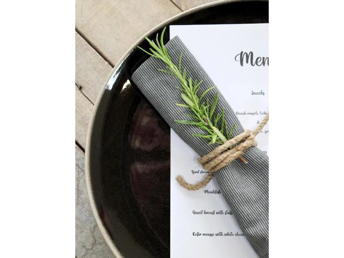 En stribet serviet bundet med sejlgarn og en kvist rosmarin hviler på en menu og en mørk tallerken.
