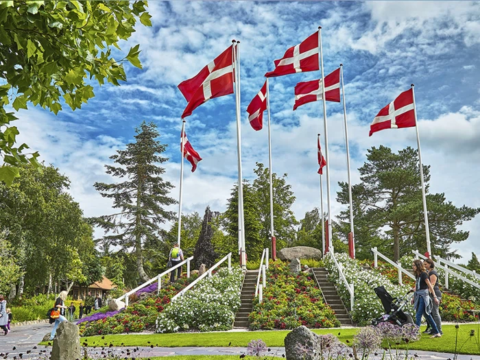 Flere danske flag på høje stænger står blandt blomsterbede og grønt i en park, og folk går rundt i nærheden under en delvist overskyet himmel.