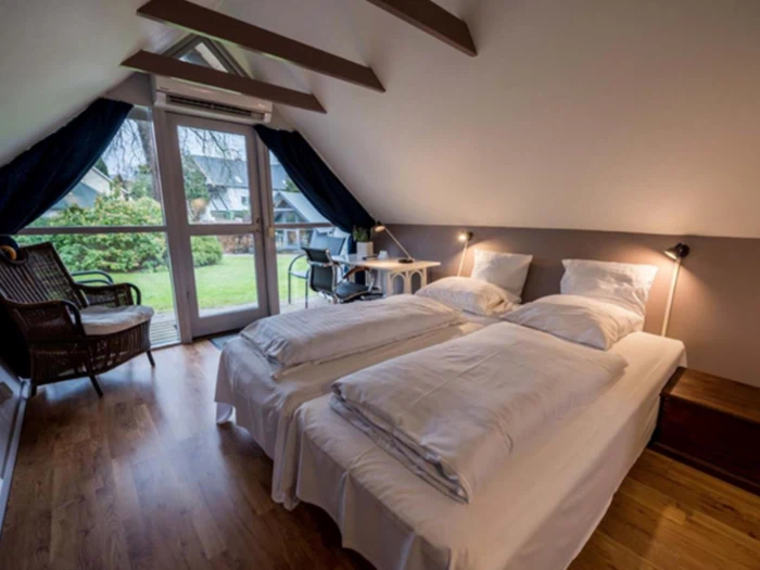 Et hyggeligt loftsværelse med to enkeltsenge, hvidt sengetøj, trægulv, skrivebord ved store vinduer og udsigt til haven udenfor.