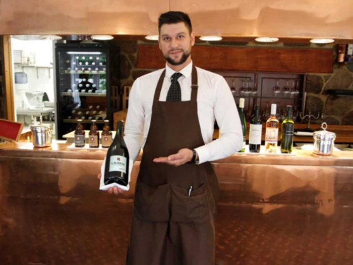 En bartender i hvid skjorte, brunt forklæde og slips står bag en bar og holder og præsenterer en flaske vin med hylder og flasker i baggrunden.