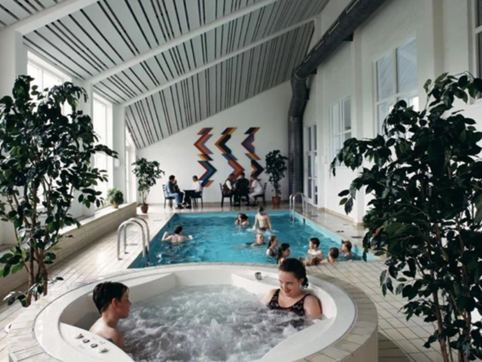 Folk slapper af i et indendørs poolområde; to sidder i et spabad i forgrunden, andre svømmer eller sidder ved poolen. Store planter og farverig vægkunst dekorerer det lyse, moderne rum.