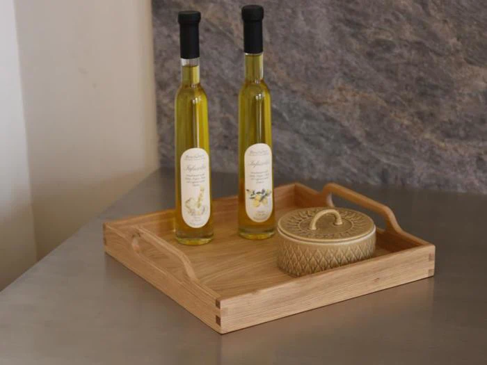 To flasker olivenolie og en dekorativ krukke med låg står på en træbakke, der er placeret på en grå bordplade.