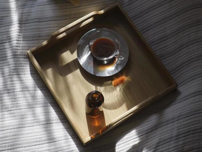 En træbakke på en stribet stofoverflade rummer en glaskop med mørk te på en underkop og en ravfarvet flaske, som begge kaster bløde skygger i det milde sollys.