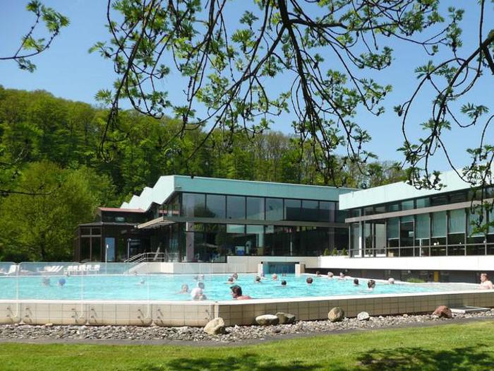 Folk svømmer og slapper af i en udendørs pool ved siden af en moderne bygning omgivet af træer og grønne områder på en solrig dag.