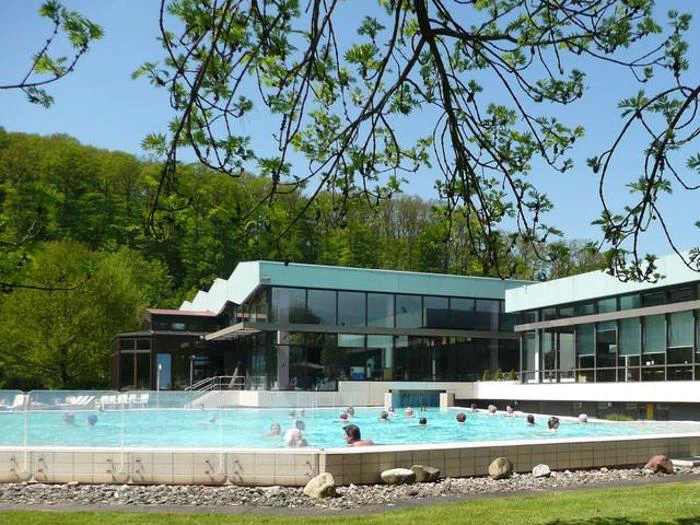 Folk svømmer og slapper af i en udendørs pool ved siden af en moderne glasbygning, omgivet af grønne træer under en klar blå himmel.