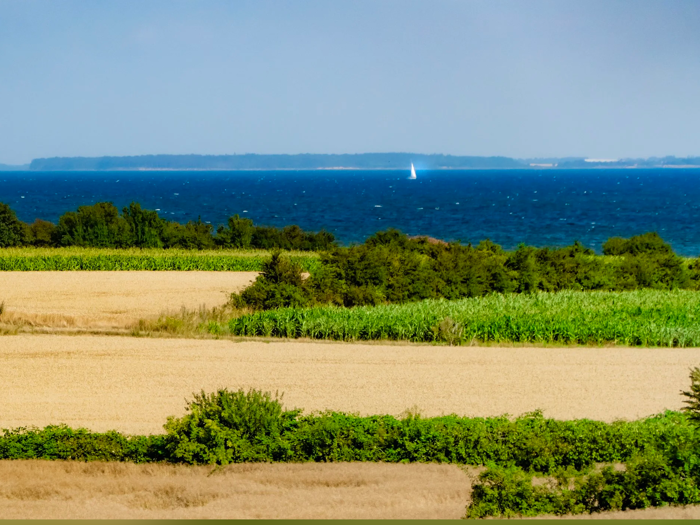 En naturskøn udsigt over marker med grønne og gyldne afgrøder, træer og et blåt hav i det fjerne. En lille hvid sejlbåd er synlig på vandet under en klar himmel.