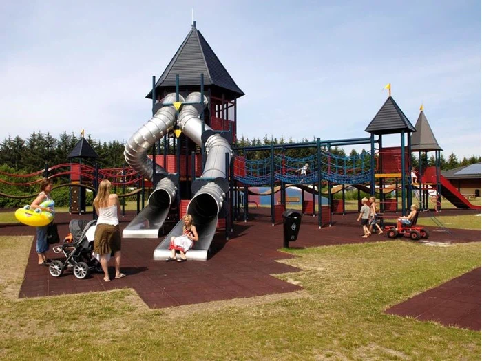 Børn og voksne leger på en udendørs legeplads med rutsjebaner, klatrereb og tårne på en solrig dag. Nogle børn er på rutsjebanerne, mens andre leger i nærheden.