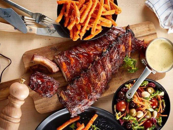 Grillede ribben på et træbræt med søde kartoffelpommes frites, en skål blandet salat og en lille gryde med cremet dippingsauce ved siden af.