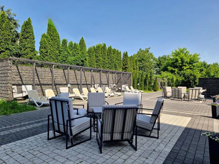 Udendørs loungeområde med stribede, polstrede stole omkring små borde, liggestole under en pergola og træer i baggrunden på en solrig dag.