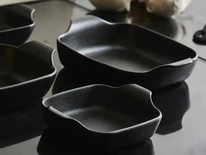 Flere sorte rektangulære keramiske bageplader i forskellige størrelser er anbragt på en mørk, reflekterende overflade.