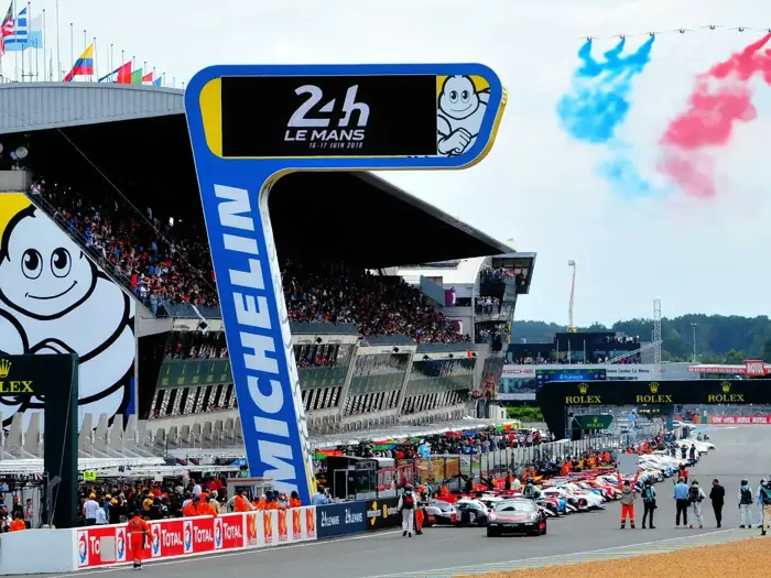 Bilerne holder i kø ved starten på 24-timers løbet i Le Mans, med tilskuere på tribunerne og rød, hvid og blå røg på himlen over et stort Michelin-skilt.
