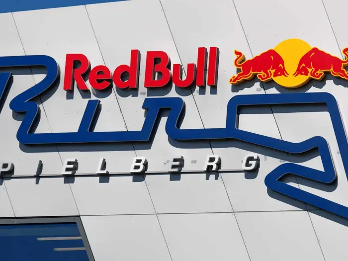 Red Bull-logo over en stiliseret blå racerbane med ordet "Spielberg" nedenunder, vist på en hvid væg.