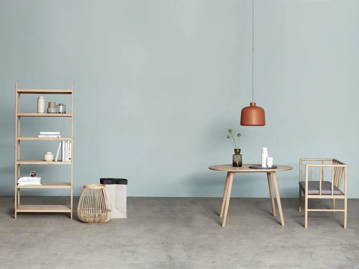 Minimalistisk rum med lyse træmøbler, bl.a. en bogreol, et bord og en stol. En brun pendel hænger over bordet, der er dækket med vaser og en plante. Lyseblå væg og gråt gulv.