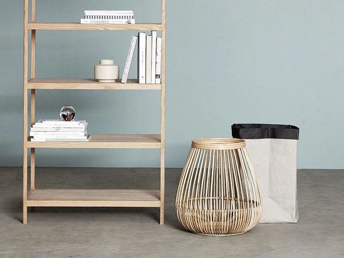 En træhylde med bøger og dekorationer står ved siden af en flettet kurv og en papirkurv mod en lysegrå væg og et betongulv.