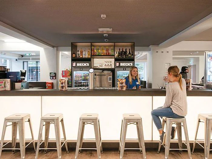 En kvinde sidder ved en moderne bardisk med hvide skamler og taler med bartenderen i et lyst, moderne rum.