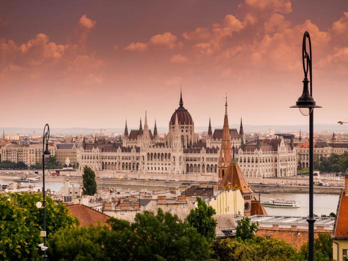 Den ungarske parlamentsbygning står ved siden af Donau-floden ved solnedgang, med dramatiske skyer på himlen og gadelamper, der indrammer bybilledet.