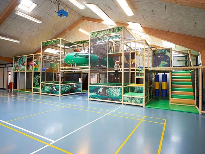 Indendørs legeplads med klatrestrukturer, rutsjebaner og bløde legeområder, omgivet af sikkerhedsnet; børn leger inde i de farverige legeredskaber.