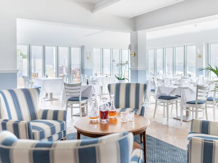 Lys, luftig restaurant med stribede lænestole og et rundt bord med glaskarafler; hvide borde og stole er arrangeret ved store vinduer i baggrunden.