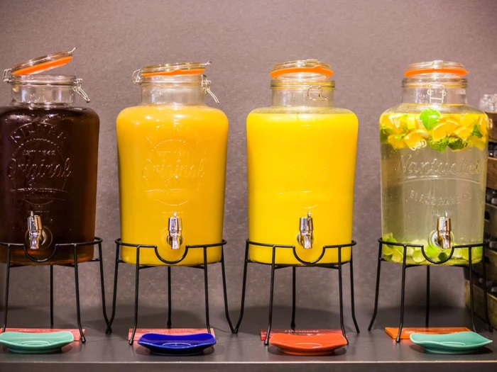 Fire glasdispensere med metalstativ, fyldt med forskellige drinks: mørkebrun, orange, gul og klar med lime- og appelsinskiver. Farvede bordskånere er placeret under hver tud.