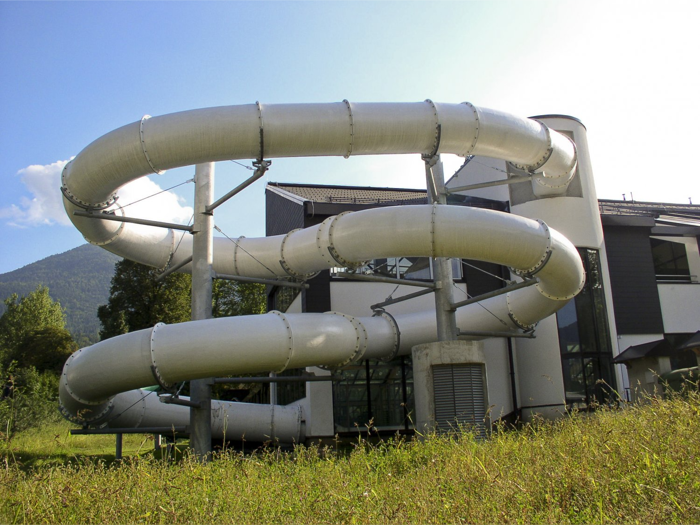 En stor, sølvfarvet, spiralformet rutsjebane snor sig rundt om en moderne bygning med græsbevoksning og træer i forgrunden.