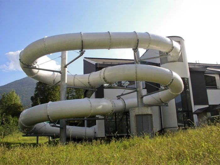 En stor, sølvfarvet, spiralformet rutsjebane snor sig rundt om en moderne bygning med græsbevoksning og træer i forgrunden.