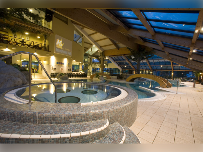 Indendørs spa med runde boblebade, en gangbro og liggestole, alt sammen under et glastag med træbjælker og varm belysning.