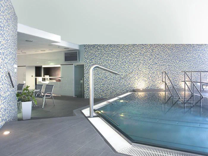 Moderne indendørs swimmingpool med vægge af mosaikfliser, gelændere i rustfrit stål og et lille opholdsområde med stole og planter i baggrunden.