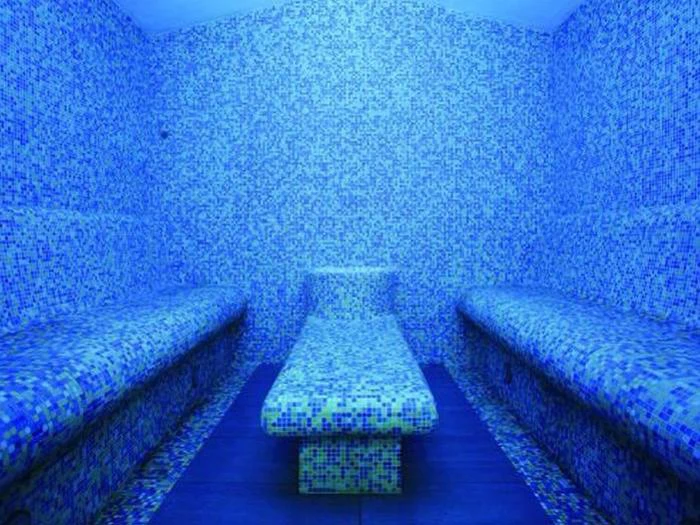 Et flisebelagt dampbad med blå mosaikvægge og bænke på begge sider samt en rektangulær platform i midten. Rummet er stærkt oplyst med en blå nuance.