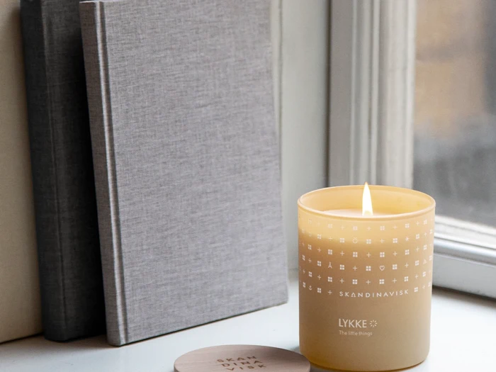 Et tændt gult lys med påskriften LYKKE står i vindueskarmen ved siden af to grå, indbundne bøger og en lysestage af træ.