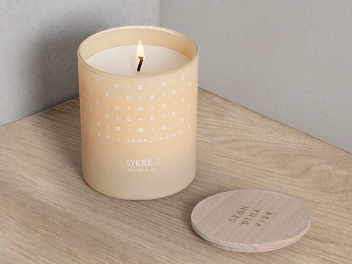Et tændt SKANDINAVISK LYKKE-lys i en cremefarvet glaskrukke står på en træplade ved siden af sit trælåg.